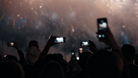 Many people's hands hold smartphones and shoot videos of exploding multi-colored fireworks in the night sky.