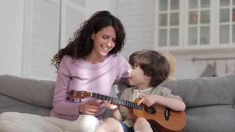Smiling mother musician teaching preschool boy to hold and play ukulele sitting on couch in living room during quarantine lockdown. Mum enjoy free time with growing child has fun together with kid