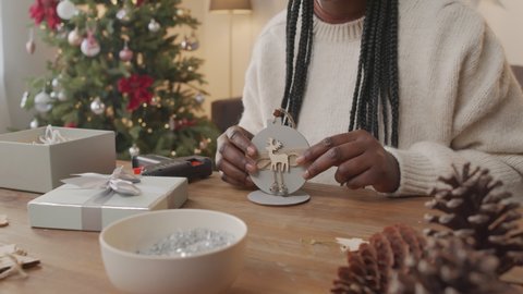 Handheld mid-section of unrecognizable African-American woman sitting at table in living room with Christmas tree and showing handmade wooden decoration to camera