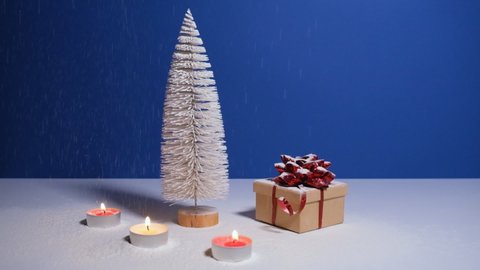 Beautiful Christmas or New Year video banner with copy space. Toy Christmas tree, gift box with red bow and burning candles on blue background with snow falling on the background