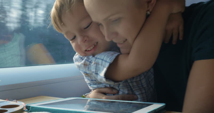 Little boy and his mother are traveling by train, tablet PC is lying on the table in front of them. Boy is hugging mother, she kisses him.