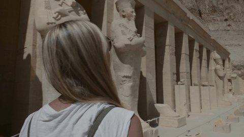 Young woman sightseeing ancient Egyptian temple in Luxor. Tracking shot: Woman walking pass Pharaoh status on vacation 