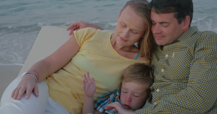 Young family relaxing on the chaise longue on the sea shore. Father and son playing on cell phone while mother stroking boys head with distracted look