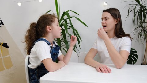 Caring mother practicing foreign language words pronunciation with small daughter in living room. Concentrated young professional speech female therapist teaching little child girl with voice problems