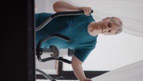 Vertical video: Elder person training on electronic stationary bicycle to do physical exercise and activity. Senior woman using cardio cycling machine to train legs muscles with gymnastics at home.