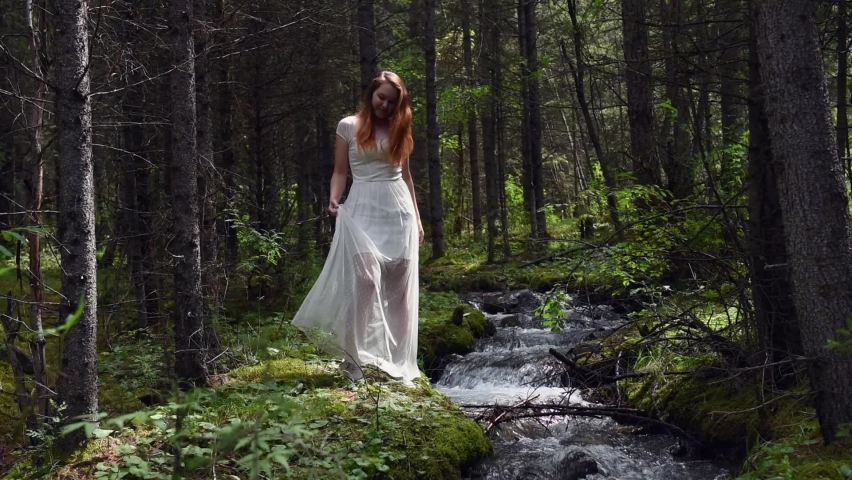 Pretty long-haired girl in white dress standing near mountain stream in dense moist forest among green trees. Happy woman enjoying outdoor recreation.