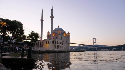 Ortakoy Mosque at Bosporus waterfront in morning hour, beautiful building illuminated from night time. Small waves on water, 15 July Martyrs Bridge seen on background.