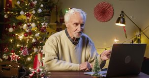 Old man pensioner speaking on video call using laptop, holding sparkler. Grandfather talking online and smiling, sitting in decorated living room. Christmas holidays during coronavirus lockdown.
