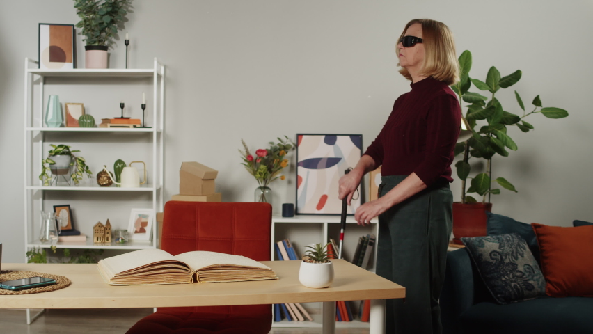 Blind woman reading braille book using fingers, sitting in living room, poorly seeing female person in glasses learning to read, home education for people with disabilities, touching letters on paper.