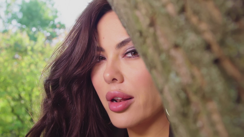 Close up of attractive curly hair brunette woman with brown eyes hiding behind tree in park at fall on sunny day. Face of beautiful woman smiling in autumn outdoor. Joy of life, fashion blogger.