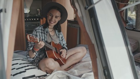 Medium shot of happy young woman in hat sitting on bed in cozy camper and playing music on ukulele