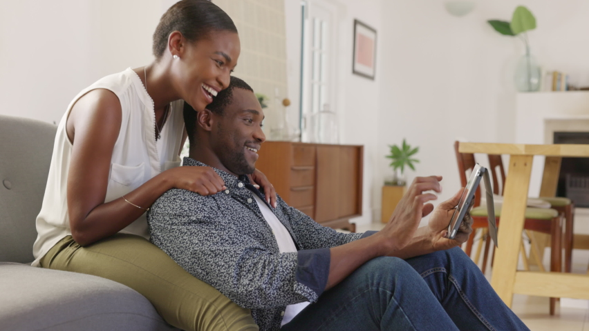 Middle aged black couple using digital tablet while sitting on couch to browse the internet surfing the net. Happy smiling black woman embracing boyfriend from behind while watching video on tablet.