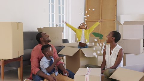 Happy black little girl playing with polystyrene foam chips while standing inside a large box. Happy african american family enjoying new home. Mature parents with son and funny daughter having fun.