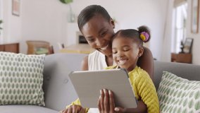 African american mature mother and little girl using digital tablet while making video call and waving hello. Happy black woman and her cute little kid sitting on sofa and doing video conference.