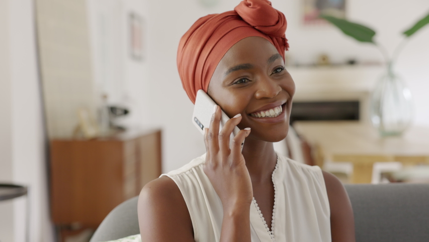 Smiling middle aged woman sitting on couch talking over smartphone. Happy black woman with traditional head turban using mobile phone for conversation. Relaxing mid african american lady using phone. Royalty-Free Stock Footage #1083153874