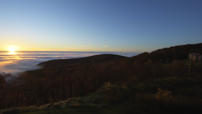 High angle aerial view above inversion clouds at sunrise morning with sun path reflection in Wintergreen Resort, Virginia ski town with colorful yellow sky and autumn trees in Blue Ridge mountains