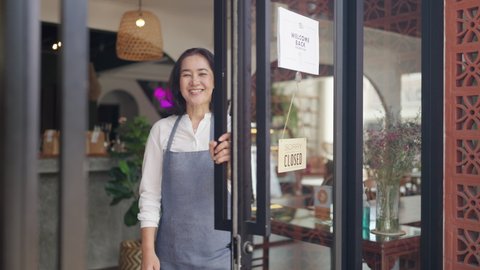 Asia latin adult female SME worker stand pride at front door work smile laugh look camera in new store small coffee shop. Happy casual day enter pub bar cafe warm hand sign for food drink industry.