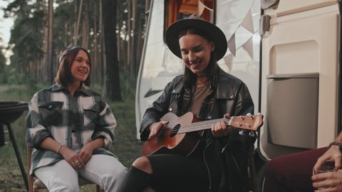 Handheld slowmo of young woman sitting before campervan parked in forest and playing ukulele while her cheerful friends singing song and enjoying music