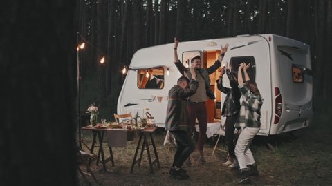 Slowmo shot of happy young people dancing together at cozy campground on summer evening. White campervan parked in background