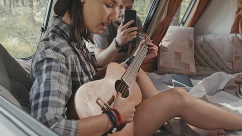 Slowmo close up of young hipster woman sitting on bed in camper and playing ukulele while her boyfriend filming her on mobile phone
