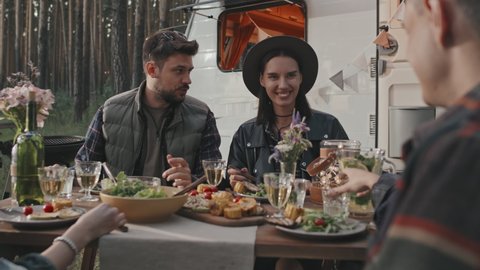 Handheld slowmo of happy young people sitting at table at campground and having dinner on summer evening. White campervan parked in background