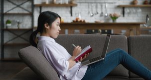 Asian woman learning distantly using laptop computer and writes notes while resting on sofa at cozy home. Female student using computer for video calling and watching online lessons on couch at home