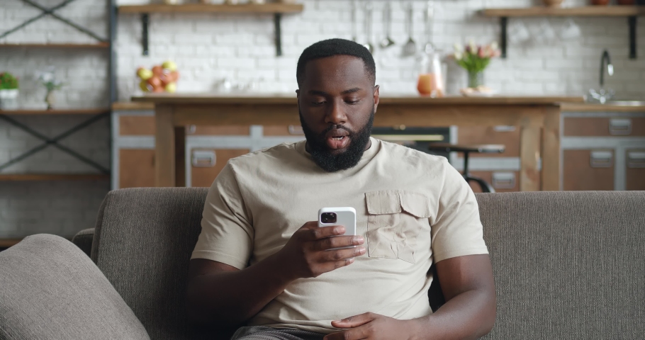 Upset black man using smartphone, loses, dissatisfied with bad result at home. Shocked african male sits on couch at home, holds mobile phone in hands on background kitchen. Royalty-Free Stock Footage #1083169882
