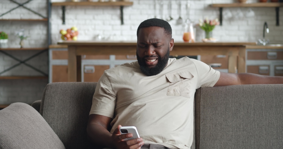 Upset black man using smartphone, loses, dissatisfied with bad result at home. Shocked african male sits on couch at home, holds mobile phone in hands on background kitchen. | Shutterstock HD Video #1083169882
