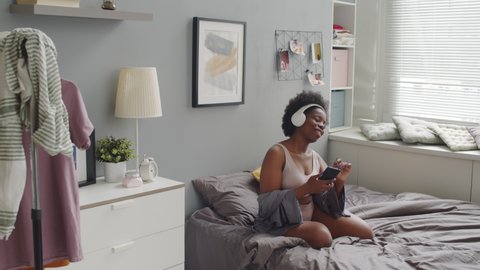 Slowmo shot of young plus size African-American woman in casual underwear enjoying music in headphones while sitting on soft bed at her bright cozy apartment