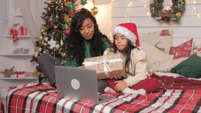 African American mother and brunette daughter in Santa hat sit on bed show present box and chat via video call using laptop slow motion