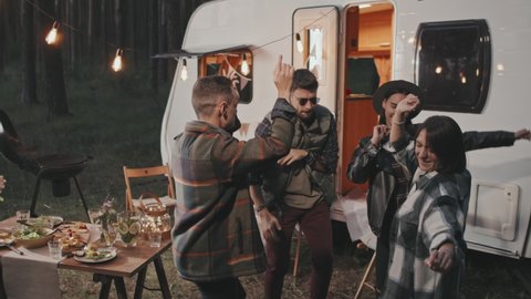 Slowmo tracking shot of young friends dancing together at campground with white camper, cozy lamp lights and picnic table laden with food. People partying on summer evening