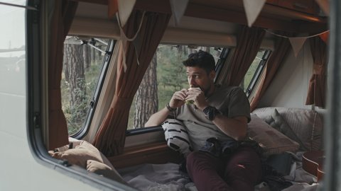 Handheld slowmo shot through window of camper parked in forest bearded young man sitting on bed and eating sandwich while enjoying nature