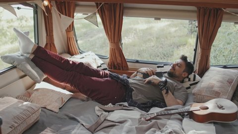 Slowmo tracking of young man relaxing on bed in cozy camper parked in forest on windy summer day
