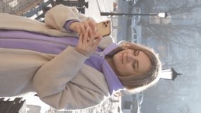 Vertical video of woman looking at phone and happy dancing. Lady in sports sweatshirt and coat using smartphone in old town