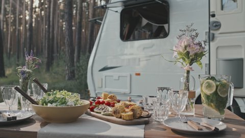 Tracking shot of tasty dishes, flowers and refreshing lemon and mint water in jug on table in forest. White camper parked in background