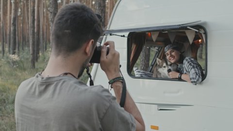 Slowmo medium shot of young man with digital camera taking photos of woman with husky looking out open window of camper parked in forest