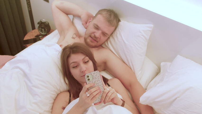 Young couple in love uses the LTE phone while lying in bed. Application for testing the compatibility of human characters. Mobile internet. Royalty-Free Stock Footage #1083177832