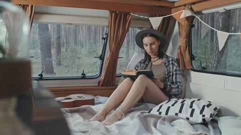 Tracking slowmo of young hipster woman in hat sitting on bed in cozy camper parked in forest and reading book
