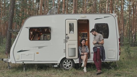 Medium shot of hipster woman in hat sitting on step in camper parked in forest and chatting with her boyfriend
