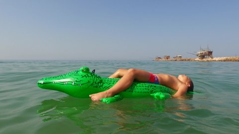 Child boy with red armband have fun laying and relaxing on floating inflatable crocodile in seawater at Punta Penna in Italy, Low-angle pov