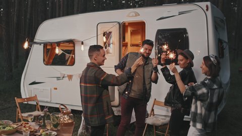 Slowmo tracking shot of happy young people with sparklers dancing at campground with cozy lights and white campervan