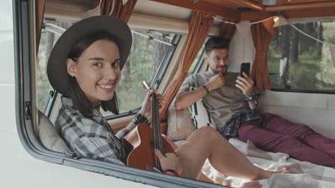 Dolly-out slowmo shot of hipster couple relaxing on bed in camper parked in forest. Young man talking on video call on mobile phone and woman playing ukulele, then looking at camera and smiling