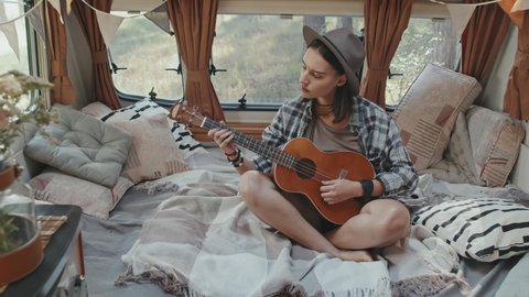 Handheld slowmo shot of young hipster woman in hat sitting on cozy bed in camper and playing music on ukulele