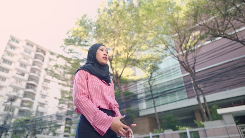 Asian female muslim wear black hijab stands on urban roadside in front of a modern glass office building with hand holding paper bags, people crossing a crosswalk, street traffic, green living space 
