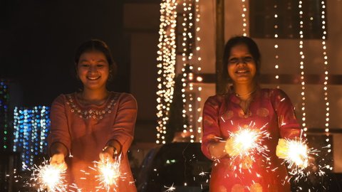 Two beautiful young Indian girls celebrating Diwali. Girls playing with fire crackers in Indian on Diwali night. Happy Indian faces on Diwali. Indian girls smiling, looking into the camera  4K