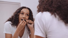 African American young female flossing her teeth with dental floss in the mirror in her modern bathroom