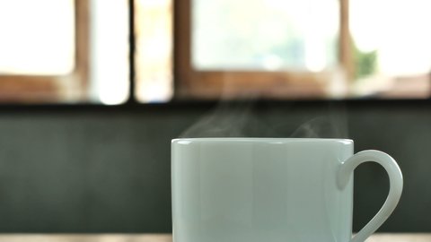 Close up white coffee cup, hot coffee, smoke rising from the coffee cup on a wooden table with shining through the window.