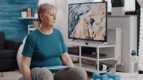 Retired person following workout video on tablet to exercise, sitting on fitness toning ball. Elder woman using elastic resistance band to stretch muscles and watching training lesson