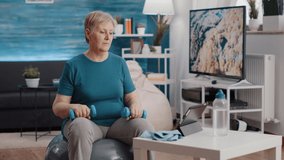 Old woman training with dumbbells to stretch arms muscles and sitting on fitness toning ball while watching online workout video on digital tablet. Senior person using weights to exercise