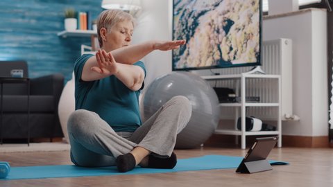 Aged person doing physical exercise and watching workout video on tablet. Senior woman following online training lesson for fitness, sitting on yoga mat. Pensioner exercising at home.
